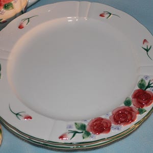 May include: White ceramic plates with a scalloped edge, adorned with red roses and green leaves. The plates are stacked, with a matching teapot and napkin rings visible. The floral design adds a touch of elegance to the tableware.