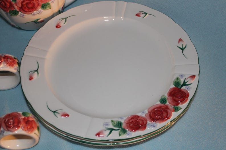 May include: White ceramic plates with a scalloped edge, adorned with red roses and green leaves. The plates are stacked, with a matching teapot and napkin rings visible. The floral design adds a touch of elegance to the tableware.