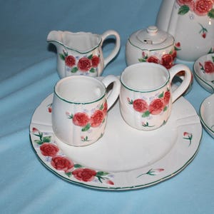 May include: A white ceramic tea set with a red rose pattern. The set includes a teapot, sugar bowl, creamer, cups, saucers, and a serving dish. The rims are trimmed with green. The background is a light blue.