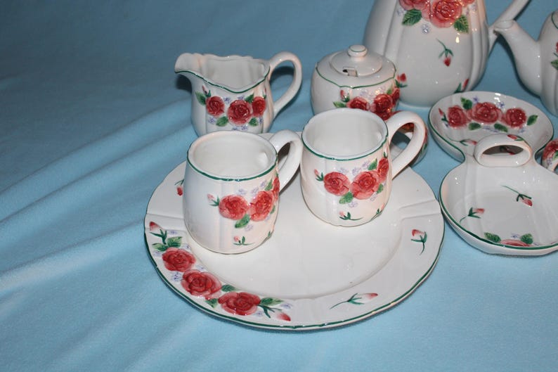May include: A white ceramic tea set with a red rose pattern. The set includes a teapot, sugar bowl, creamer, cups, saucers, and a serving dish. The rims are trimmed with green. The background is a light blue.