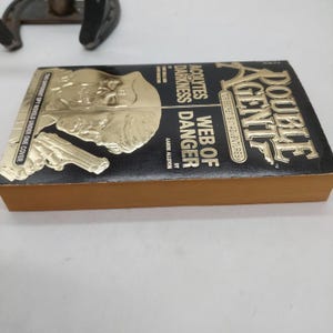 Peut inclure: Un livre de poche vintage intitul&eacute; "Double Agent: Web of Danger". La couverture est noire avec des lettres dor&eacute;es et des images en relief d'un visage d'homme et d'une arme de poing. Les pages du livre sont dor&eacute;es.