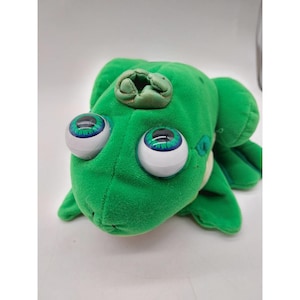 May include: A bright green plush frog toy featuring large, round, white plastic eyes with detailed blue and green irises. The frog has a small, circular, fabric-covered opening on the top of its head and a soft, velvety texture throughout its body.