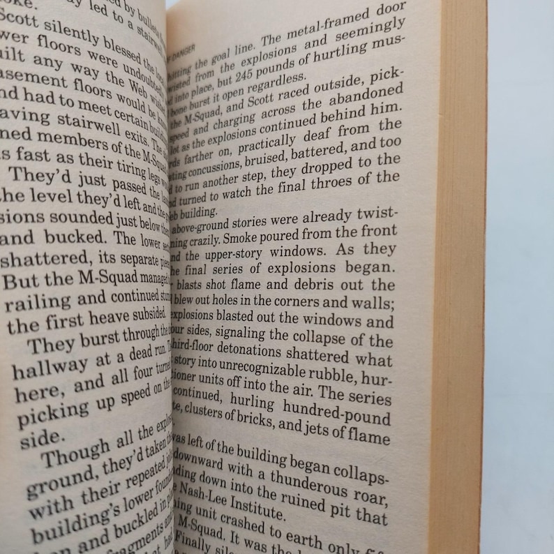 Peut inclure: Pages d'un livre ouvert avec du texte imprim&eacute;. Le texte d&eacute;crit une sc&egrave;ne avec des explosions, une escouade et un b&acirc;timent en effondrement. Les pages sont jaunies, sugg&eacute;rant l'&acirc;ge. Le livre est ouvert, r&eacute;v&eacute;lant le texte.