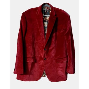 May include: A red velvet blazer with a two-button closure and a patterned interior lining. The blazer has two front pockets and a notched lapel. The jacket is hanging on a black hanger.