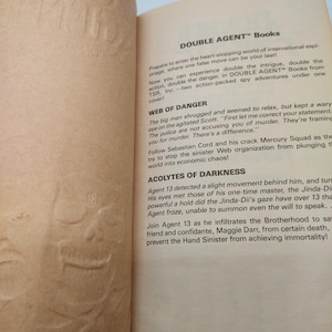 Peut inclure: Page d'un livre ouvert avec du texte sur les aventures d'espionnage "DOUBLE AGENT". Le texte comprend des titres de chapitres tels que "WEB OF DANGER" et "ACOLYTES OF DARKNESS". Le fond est textur&eacute; et blanc cass&eacute;.