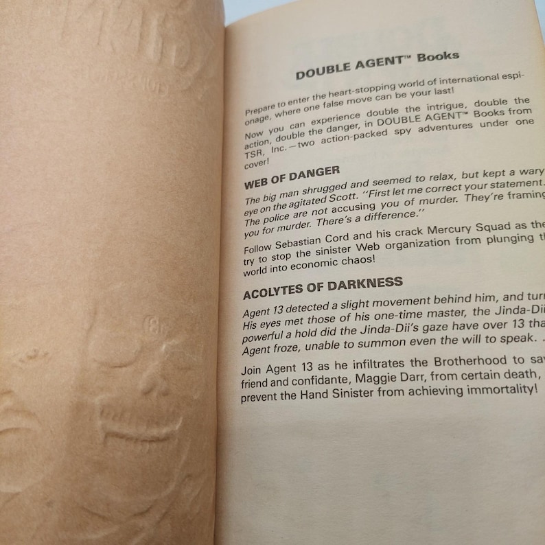 Peut inclure: Page d'un livre ouvert avec du texte sur les aventures d'espionnage "DOUBLE AGENT". Le texte comprend des titres de chapitres tels que "WEB OF DANGER" et "ACOLYTES OF DARKNESS". Le fond est textur&eacute; et blanc cass&eacute;.