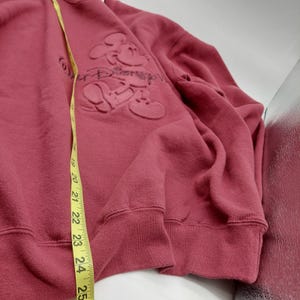 May include: A dusty rose-colored sweatshirt featuring an embossed Mickey Mouse design. The sweatshirt has a crew neck and long sleeves. A yellow measuring tape is draped over the garment, indicating its size.
