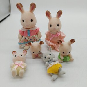 May include: A collection of six flocked animal figurines resembling rabbits. Two larger figures stand in the back wearing pink dresses, while four smaller figures in the front wear yellow, green, and white outfits. Each figure has brown-tipped ears.