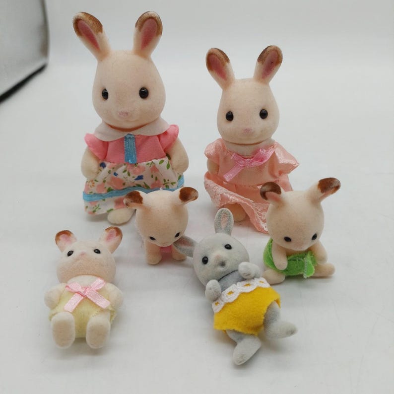 May include: A collection of six flocked animal figurines resembling rabbits. Two larger figures stand in the back wearing pink dresses, while four smaller figures in the front wear yellow, green, and white outfits. Each figure has brown-tipped ears.
