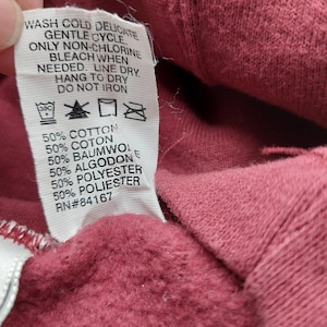 May include: A close-up of a burgundy fabric with a white tag attached. The tag displays washing instructions, including "WASH COLD DELICATE" and fabric composition details: 50% cotton, 50% polyester. The RN number is 84167.