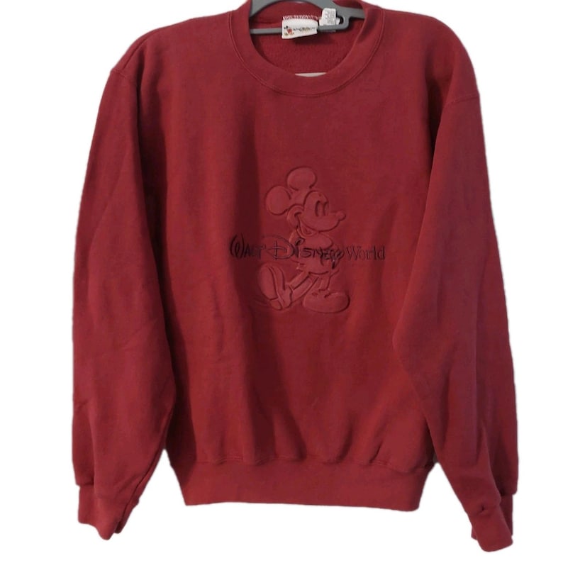 May include: A maroon crewneck sweatshirt featuring an embossed image of Mickey Mouse and the words "Walt Disney World". The sweatshirt has long sleeves and a ribbed neckline, cuffs, and hem. The Disney logo is visible on the upper back.