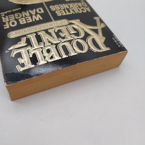 Peut inclure: Un livre &agrave; couverture rigide intitul&eacute; "Double Agent" avec des lettres dor&eacute;es sur une couverture noire. Le titre du livre est bien en &eacute;vidence, avec les mots "Web of Danger" et "Acolytes of Darkness" &eacute;galement visibles. Les pages du livre sont de couleur beige clair.