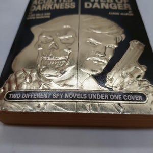 Peut inclure: Un livre &agrave; couverture rigide noire avec des illustrations dor&eacute;es en relief. La couverture pr&eacute;sente un cr&acirc;ne et un homme tenant une arme. Le texte indique "TWO DIFFERENT SPY NOVELS UNDER ONE COVER".