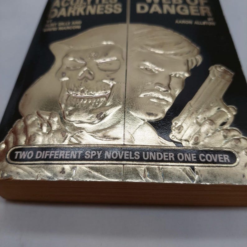 Peut inclure: Un livre &agrave; couverture rigide noire avec des illustrations dor&eacute;es en relief. La couverture pr&eacute;sente un cr&acirc;ne et un homme tenant une arme. Le texte indique "TWO DIFFERENT SPY NOVELS UNDER ONE COVER".