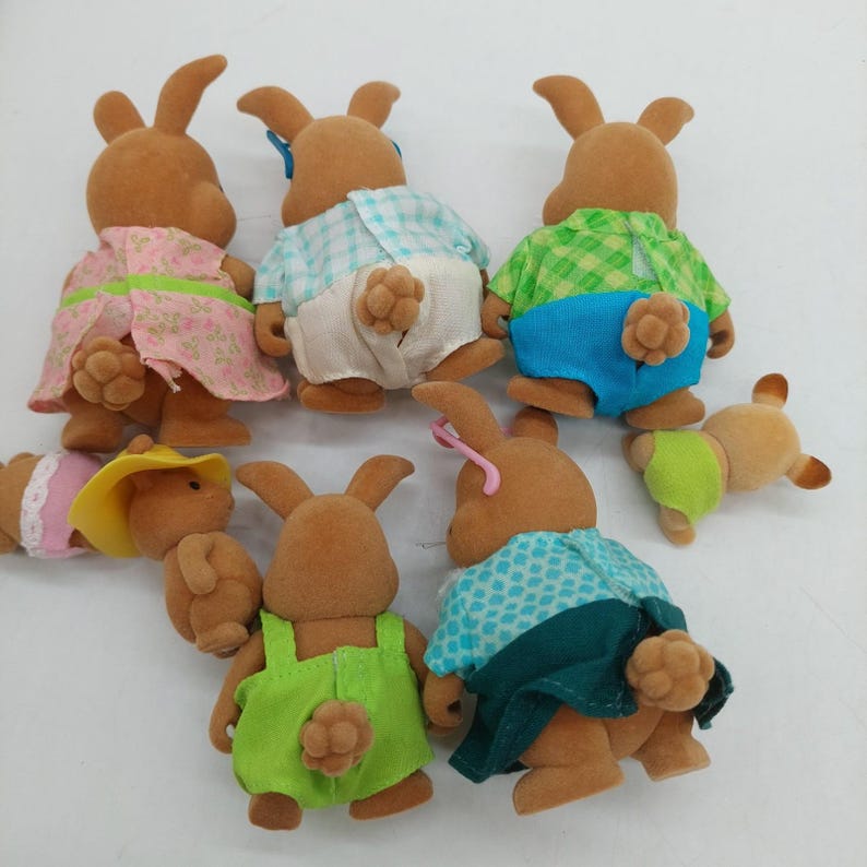 May include: A collection of seven flocked rabbit figurines viewed from the back. The figures are dressed in various colorful outfits including patterned dresses, checkered shirts, and solid-colored shorts, each featuring a small, textured tail.