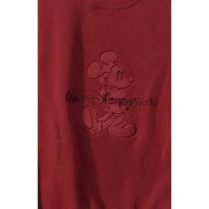 May include: A maroon sweatshirt featuring a raised design of Mickey Mouse and the text "Walt Disney World". The design is in the same color as the sweatshirt. The sweatshirt is made of a soft material.