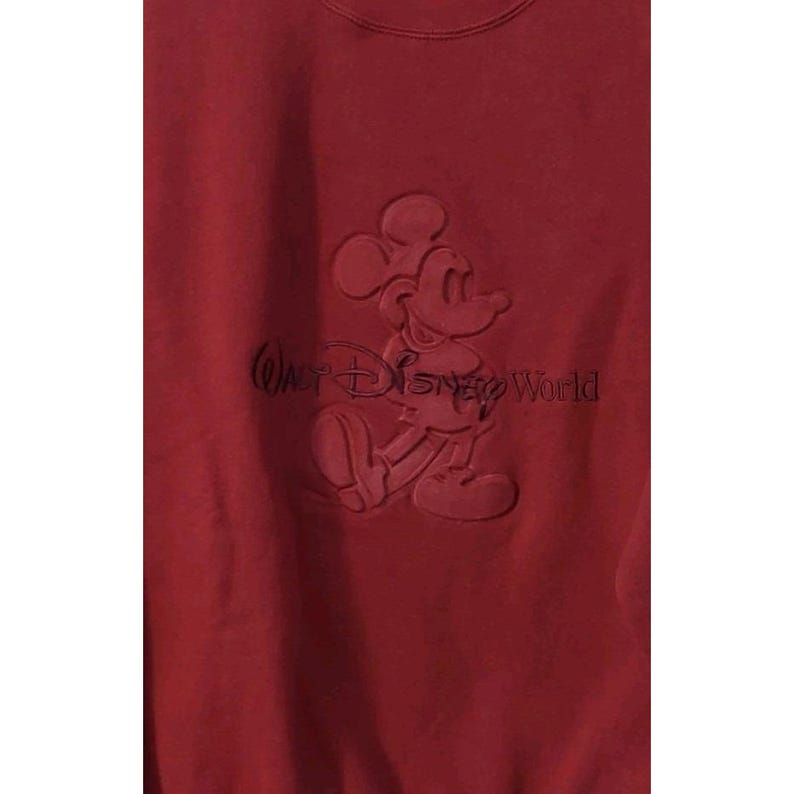 May include: A maroon sweatshirt featuring a raised design of Mickey Mouse and the text "Walt Disney World". The design is in the same color as the sweatshirt. The sweatshirt is made of a soft material.