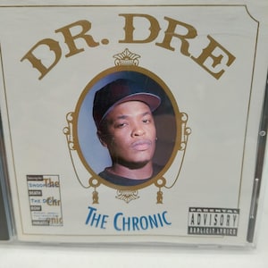 May include: A Dr. Dre The Chronic music CD case featuring a white cover with gold lettering. A central oval frame contains a portrait of a person wearing a black baseball cap. A Parental Advisory Explicit Lyrics label is in the bottom right corner.