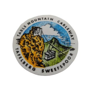 Op de afbeelding: Een ronde sticker met een grafische illustratie van de Table Mountain kabelbaan. Het ontwerp bevat een gele bergtop, een groen landschap, een blauwe lucht en een kabelbaan. De tekst op de sticker luidt TABLE MOUNTAIN CABLEWAY TAFELBERG SWEEFSPOOR.