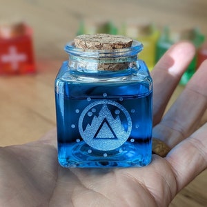 Op de afbeelding: Een klein blauw glazen flesje met een kurken stop. De fles heeft een wit bergontwerp met een cirkel van stippen eromheen.