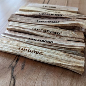 May include: A stack of light brown palo santo sticks with dark brown grain. Each stick has a different affirmation engraved in black, including "I AM LOVING," "I AM STRONG," and "I AM CENTERED."