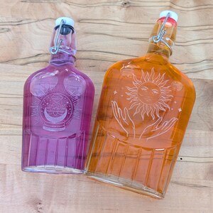 May include: Two glass bottles with swing-top stoppers. The left bottle is purple and features an etched design of a crystal ball with a lotus and crescent moon. The right bottle is orange and features an etched design of a sun above two hands.