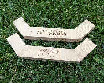 May include: Two light-colored wooden blocks with angled ends, resting on green grass. The top block has the word "ABRACADABRA" etched into it. The bottom block has a series of etched symbols. The blocks are likely decorative or functional.