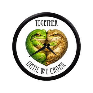 May include: Round wall clock with a black frame. The clock face features two hugging frogs, one green and one brown, forming a heart shape. The words "TOGETHER" and "UNTIL WE CROAK" are printed on the clock face.