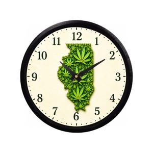 May include: A round wall clock with a black frame and a cream-colored face featuring black Arabic numerals. In the center, a map of the state of Illinois is filled with a dense pattern of green cannabis leaves. Two black clock hands are centered.