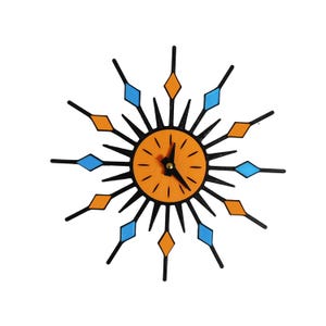 May include: A starburst wall clock featuring a central orange circular face with black hour markers and hands. The frame consists of black radiating spikes, alternating with orange and light blue diamond-shaped accents at the tips of the spikes.