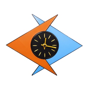 May include: A retro-style wall clock with a black clock face, gold hands, and gold hour markers. The clock is set within a geometric design of orange and blue shapes.