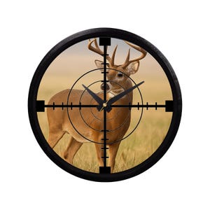 May include: A round wall clock featuring a deer in a field, framed by a black bezel. The clock face includes a crosshair design, with black clock hands indicating the time. The deer is brown with large antlers.