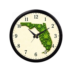May include: A round wall clock with a black frame and a cream-colored face featuring black Arabic numerals. The center displays a map of Florida filled with a repeating pattern of green cannabis leaves, accompanied by black hour, minute, and second hands.