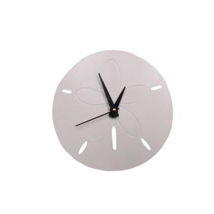 May include: A round, off-white wall clock shaped like a sand dollar. The clock features black hands and a black center. The sand dollar design has a flower-like pattern with small holes around the edges. The clock is a decorative item.