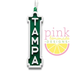 May include: A green and white ornament in the shape of a street sign, with the word "TAMPA" in white capital letters. The ornament has a white border and a green background. A string is attached for hanging.