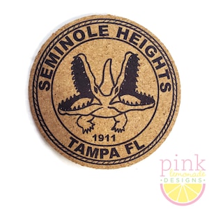 May include: Round cork coaster with a black design featuring two alligators and the text "SEMINOLE HEIGHTS TAMPA FL 1911". The coaster is a natural tan color with a textured surface.