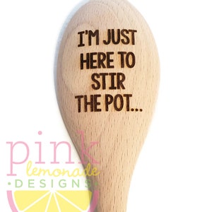 May include: Wooden spoon with the engraved text "I'M JUST HERE TO STIR THE POT...". The spoon is light brown and has a rounded end. The Pink Lemonade Designs logo is visible at the bottom.