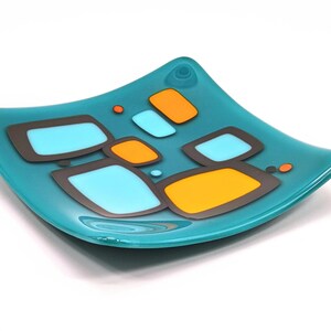 Könnte beinhalten: Eine türkisfarbene Glasschale mit quadratischer Form und geschwungenen Kanten. Die Schale hat ein geometrisches Design aus orangefarbenen, hellblauen und grauen Rechtecken. Ein dekoratives Element für das Zuhause.