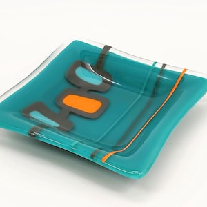 May include: A teal blue glass dish with a geometric design in orange and grey. The dish has a slightly curved shape and a smooth, glossy finish.