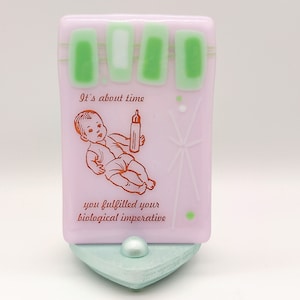 May include: A pink glass tile with a green and white design. The tile has a picture of a baby holding a bottle with the text "It's about time you fulfilled your biological imperative".
