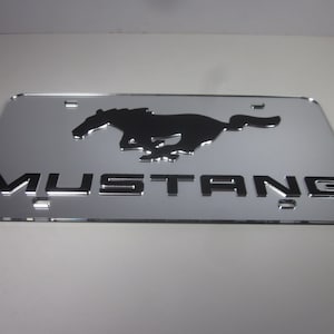 May include: A silver metal license plate with a black silhouette of a running horse and the word "MUSTANG" in black lettering.