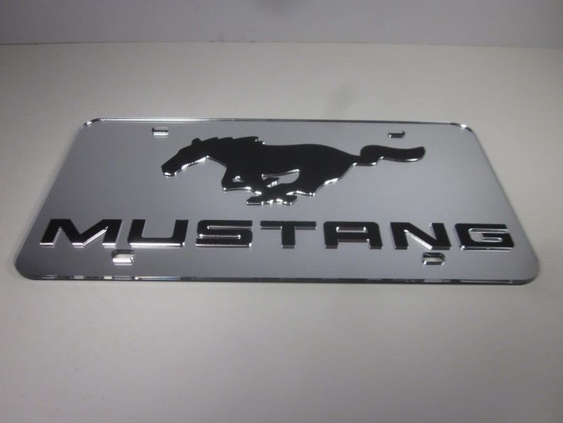 Puede incluir: Una placa de matr&iacute;cula de metal plateado con una silueta negra de un caballo corriendo y la palabra "MUSTANG" en letras negras.