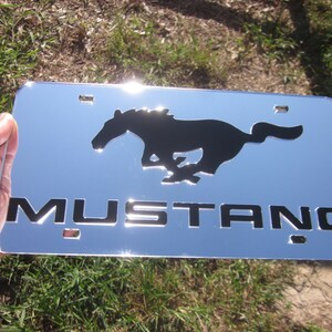 Puede incluir: Marco de matr&iacute;cula cromado con una silueta negra de un caballo corriendo y la palabra "MUSTANG" en letras negras.