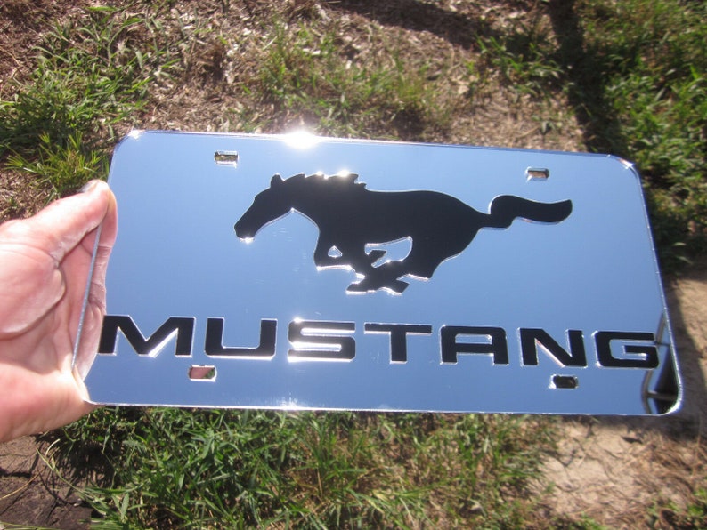 Puede incluir: Marco de matr&iacute;cula cromado con una silueta negra de un caballo corriendo y la palabra "MUSTANG" en letras negras.