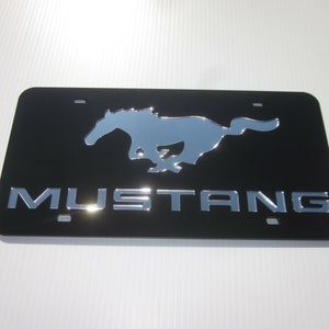 Puede incluir: Marco de matr&iacute;cula de metal negro con un logotipo de Mustang en cromo plateado y la palabra "MUSTANG" en letras de cromo plateado.
