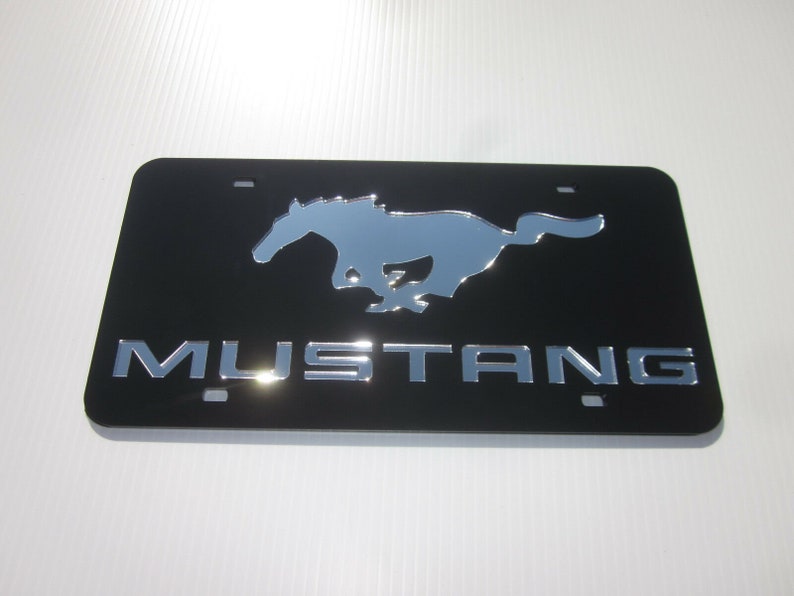Puede incluir: Marco de matr&iacute;cula de metal negro con un logotipo de Mustang en cromo plateado y la palabra "MUSTANG" en letras de cromo plateado.