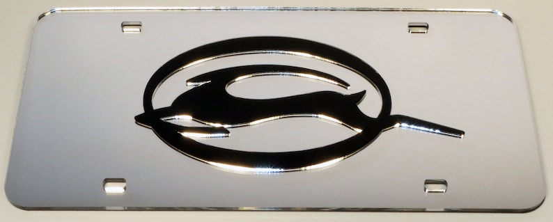 May include: A silver chrome license plate frame with a raised, three-dimensional, black Chevrolet Bowtie logo. The logo features a stylized running deer inside a circle.