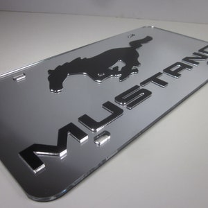 Puede incluir: Una placa de matr&iacute;cula cromada plateada con una silueta negra de un caballo corriendo y la palabra "MUSTANG" en letras en relieve.