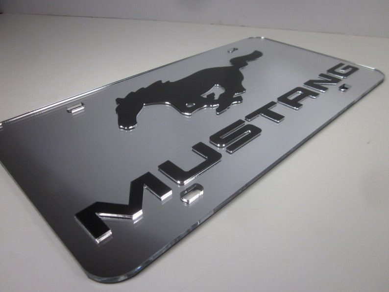 Puede incluir: Una placa de matr&iacute;cula cromada plateada con una silueta negra de un caballo corriendo y la palabra "MUSTANG" en letras en relieve.