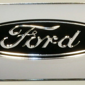 Può includere: Un telaio per targa argentato con un ovale nero che presenta la parola "Ford" in un font script argentato.